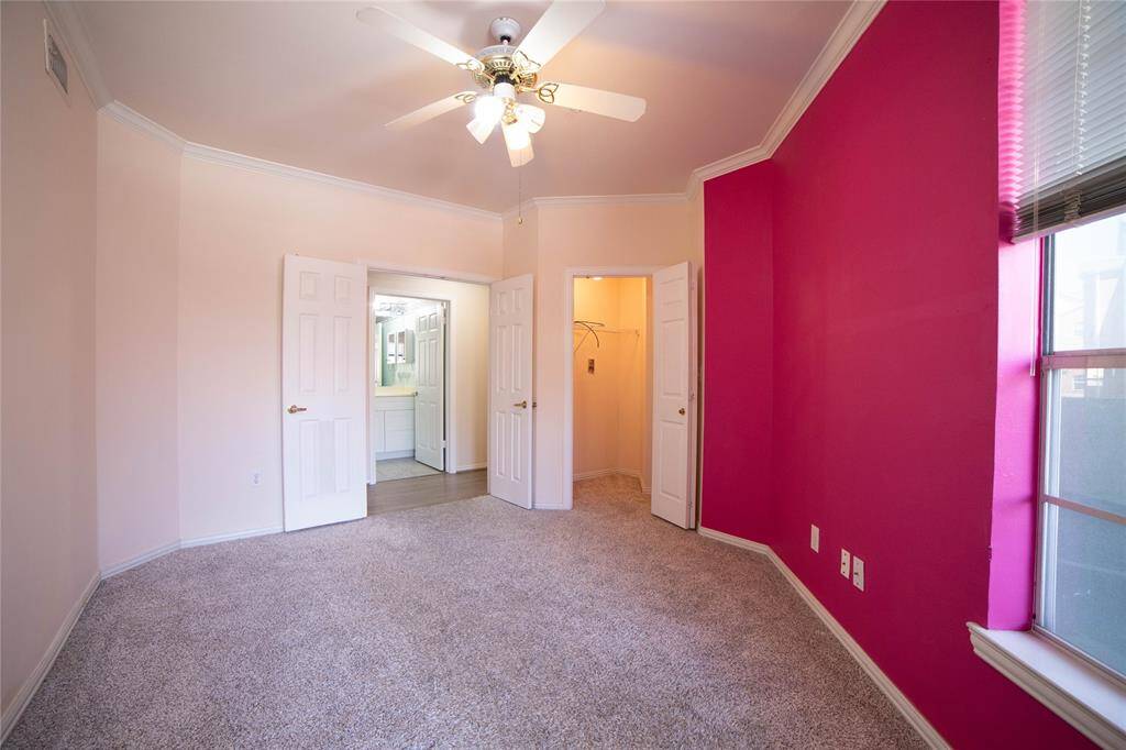 Bright bedroom with plush carpeting and a bold pink accent wall. Features a ceiling fan, ample natural light from a large window, and access to a walk-in closet and bathroom.