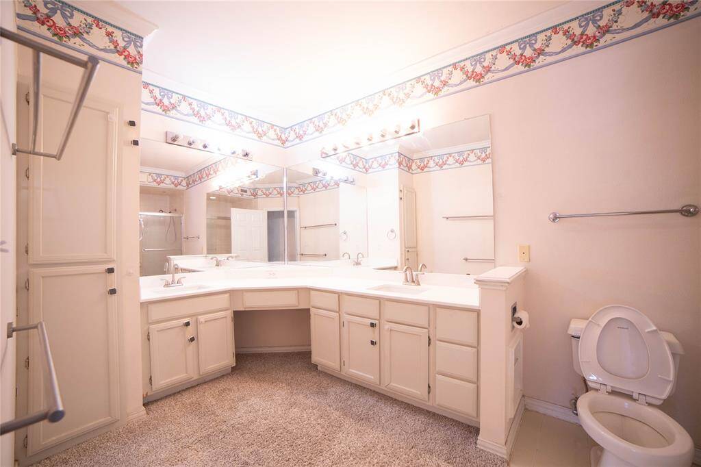 A spacious bathroom with dual sinks, ample cabinet storage, large mirrors, and decorative wallpaper border. It has a cozy carpeted floor and bright lighting, creating a welcoming space.