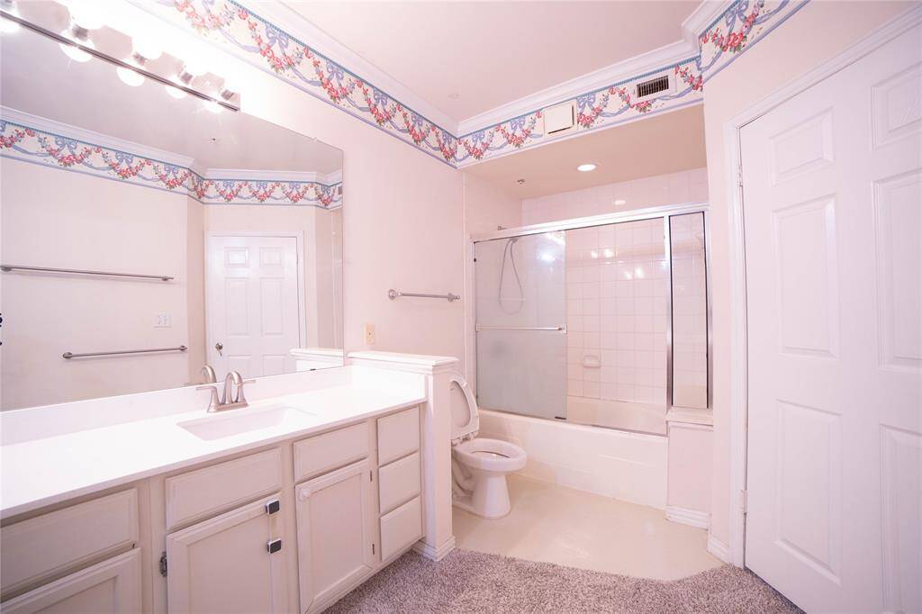 This bathroom features a large vanity with ample storage, a full-width mirror, and a bathtub with a sliding glass door. The room is accented with floral wallpaper trim and has a soft carpeted floor.