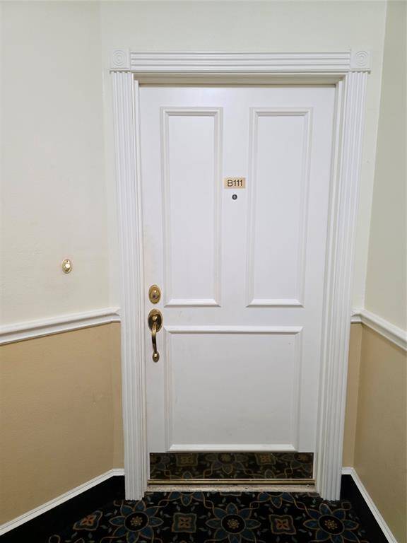 This photo shows a white, paneled door with a brass handle and a peephole, framed by decorative molding. It has a unit number "B111" and is set against a two-tone wall with a patterned carpet below.