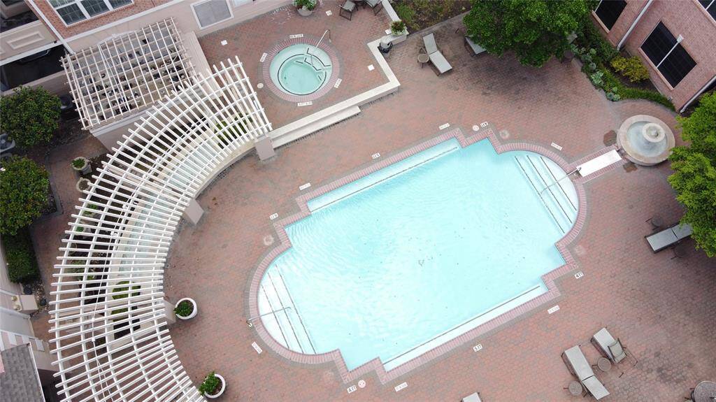 This photo showcases a communal outdoor area featuring a large, stylish swimming pool with an adjacent hot tub. The space is surrounded by a brick patio with lounge chairs, a decorative pergola, and lush greenery, offering a relaxing environment for residents.