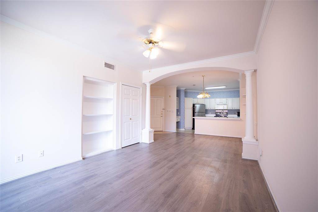 This photo showcases a spacious, open-concept living area with light wood flooring and neutral walls. It features built-in shelving, a ceiling fan, and a view into a modern kitchen with stainless steel appliances. Elegant columns frame the transition between the living space and the kitchen.