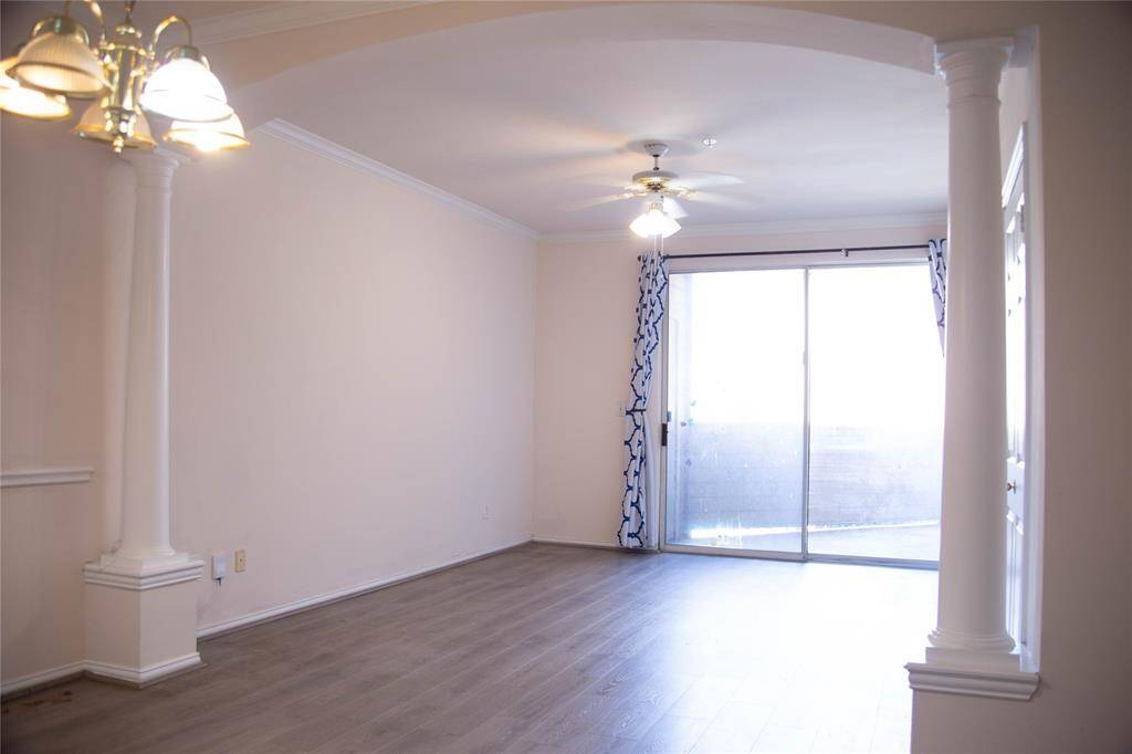 This photo shows a bright, open living space with hardwood floors, a ceiling fan, and a large sliding glass door leading to a balcony. Decorative columns and a chandelier enhance the room's elegance.