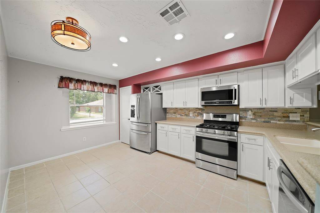 This modern kitchen features stainless steel appliances, white cabinetry, and a tile floor. A  window provides natural light, complemented by recessed lighting and a stylish ceiling fixture. The room is accented with red trim and a stone backsplash.