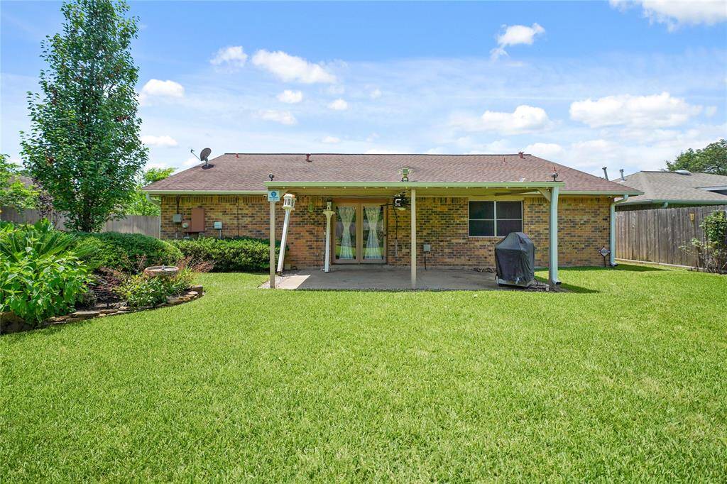 Photo shows the backyard of a brick house featuring a covered patio area. The yard is spacious and well-maintained, with green grass and surrounding landscaping, offering a private and inviting outdoor space for relaxing or entertaining.