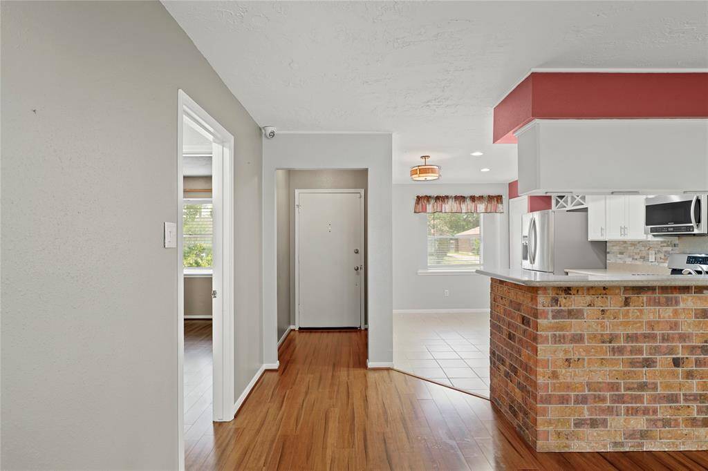 Photo showcases a welcoming entryway leading to a kitchen that opens to the living area. The space features vinyl wood flooring, a brick accented wall on the kitchen counter, modern stainless steel appliances, and ample natural light from large windows.