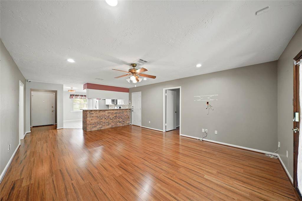 This photo showcases a spacious open-concept living area with vinyl wood flooring. A ceiling fan is centrally located, and the room connects to a kitchen with a brick accent and modern appliances, offering a welcoming and versatile space.