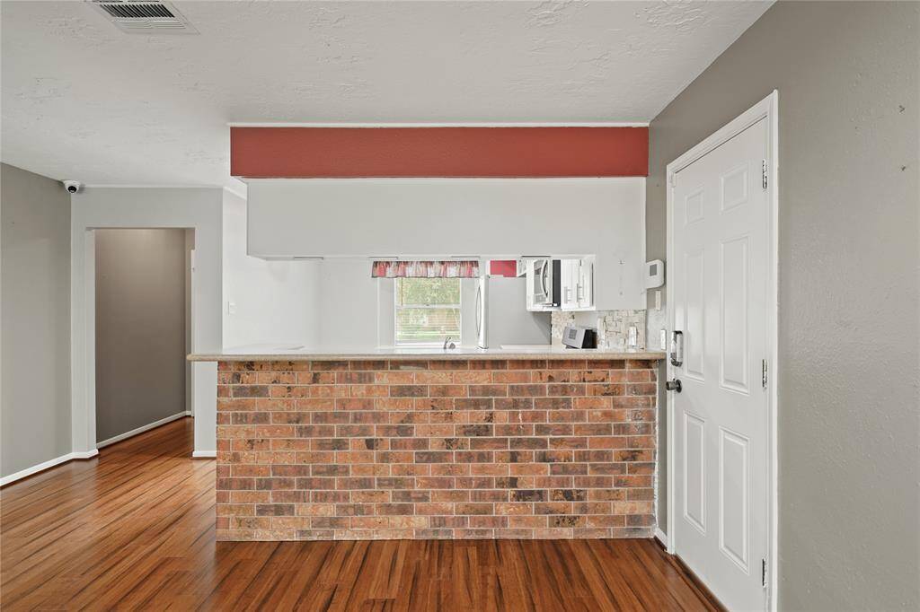 This photo shows a view of the  cozy kitchen area through a brick accented counter opening from the living area. The kitchen is partially visible, featuring stainless steel appliances and a window with a view. The layout is open and inviting.