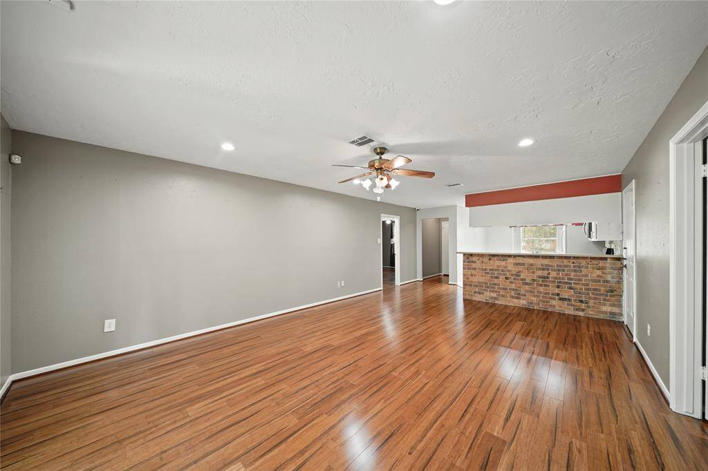 This spacious living area features hardwood vinyl floors and recessed lighting. A brick accent wall adds character, and a ceiling fan provides comfort. The open layout connects to the kitchen, creating a welcoming space.