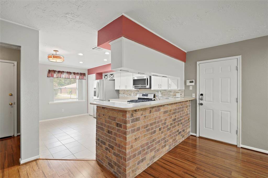 This photo showcases a bright, open kitchen with modern appliances and white cabinetry. The space features a brick accent bar with tile flooring in the kitchen and recessed lightening. Door pictured leads to the 2 car garage.