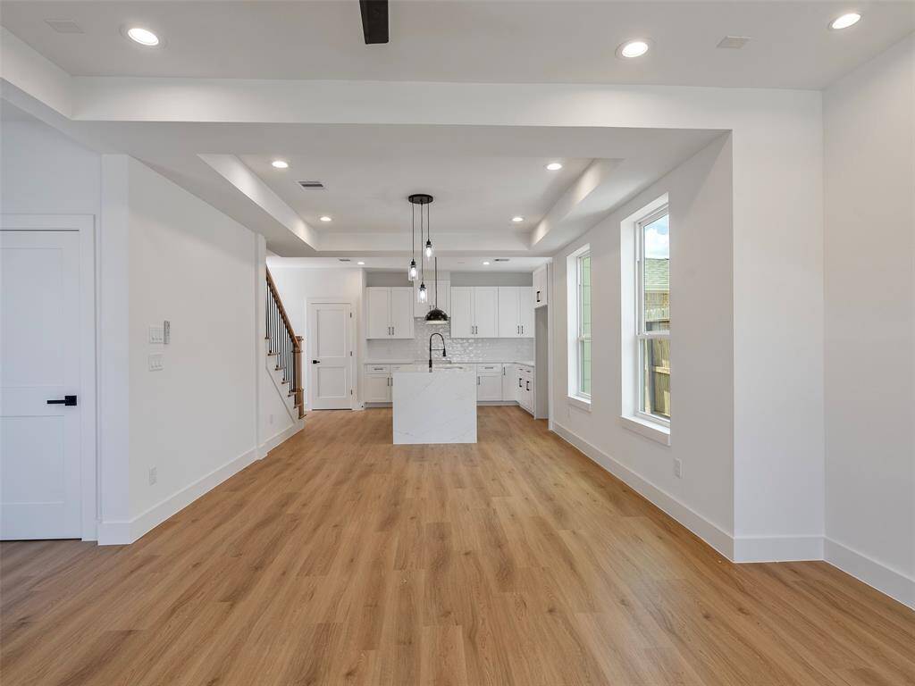 Modern, open-concept kitchen and living area with hardwood floors, recessed lighting, and large windows for natural light. The kitchen features white cabinetry, a central island with a sink, and stylish pendant lights.