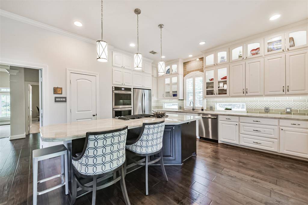 This kitchen features modern lighting, a spacious island with seating, stainless steel appliances, and ample white cabinetry for storage, all set against elegant dark wood flooring.