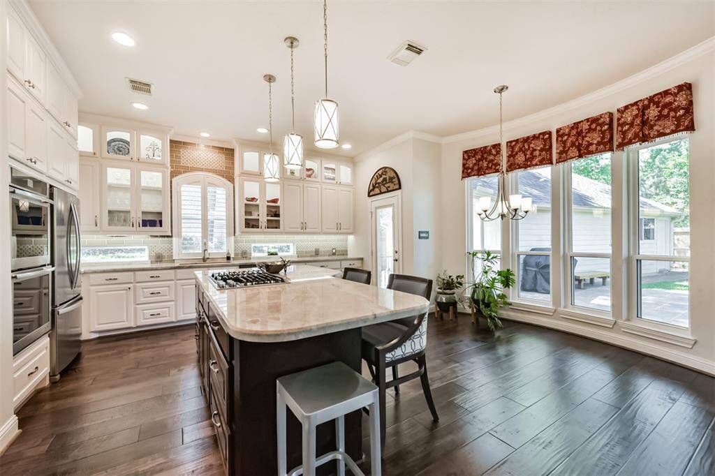 This spacious kitchen features elegant white cabinetry, a large center island with seating, and stainless steel appliances. It boasts ample natural light from expansive windows and offers a cozy dining area with stylish pendant lighting. Perfect for both cooking and entertaining.