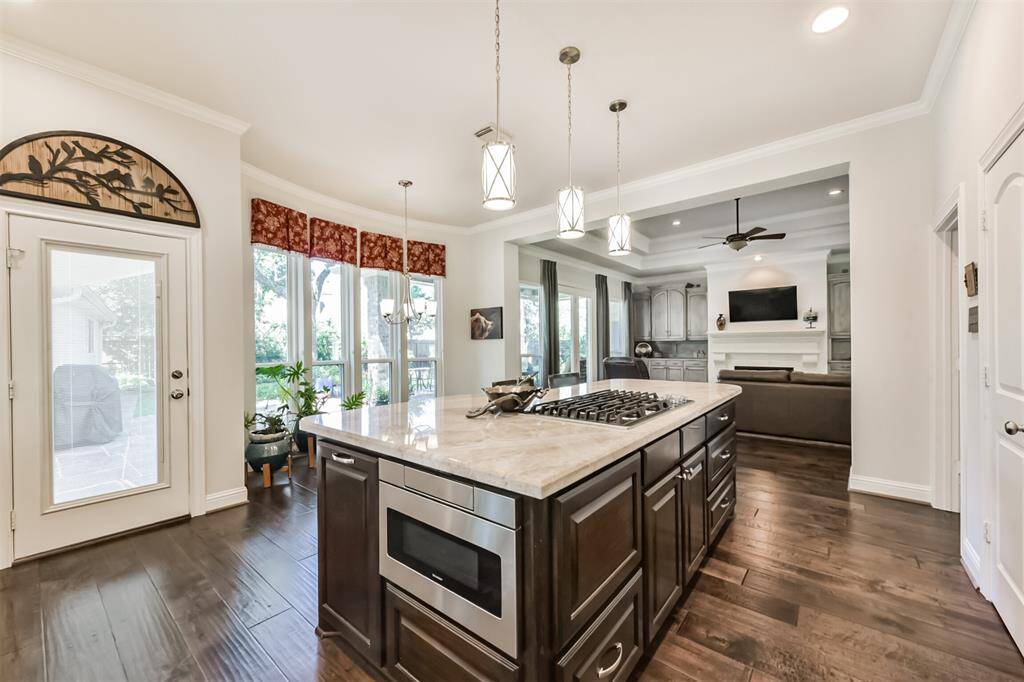 This spacious kitchen features a large island with a built-in stove, modern pendant lighting, and elegant dark wood cabinetry. It opens to a bright living area with large windows, hardwood floors, and a cozy fireplace, perfect for entertaining and family gatherings.