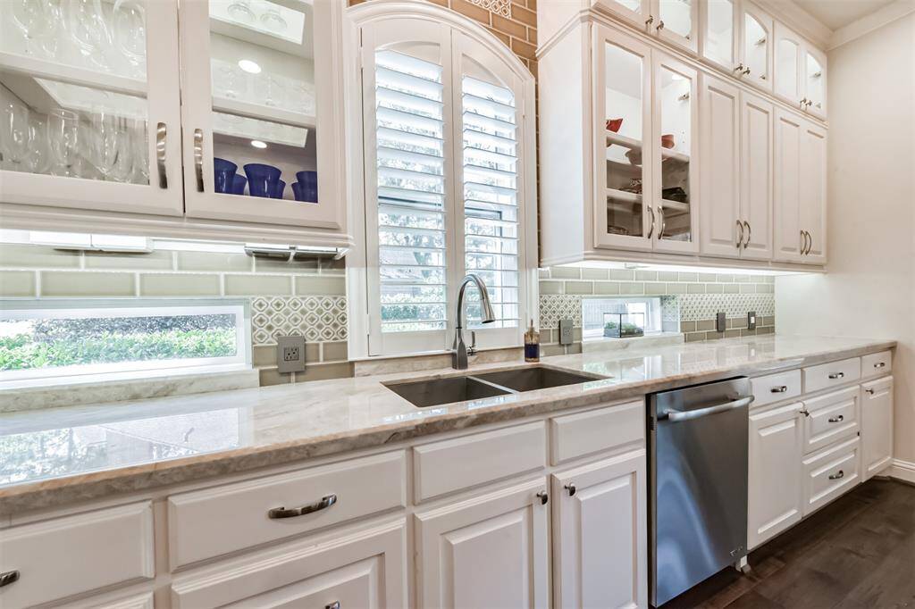 This kitchen features elegant white cabinetry with glass-front doors, a sleek marble countertop, and a modern stainless steel dishwasher. The space is well-lit with natural light from a window with plantation shutters, and has stylish tile backsplash accents.