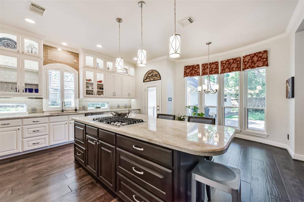 This spacious kitchen features elegant cabinetry with glass display sections, a large central island with a gas cooktop, and modern pendant lighting. It offers ample natural light through large windows, complementing the open and airy design. The dark wood flooring contrasts beautifully with the light-colored countertops and cabinets, creating a warm and inviting atmosphere.