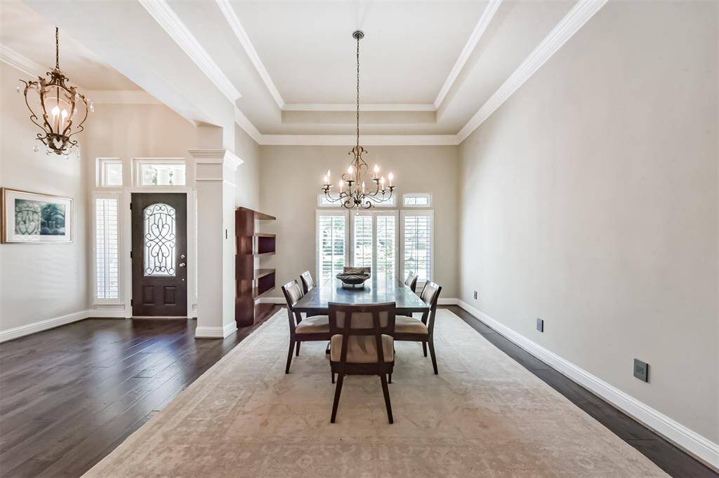 This elegant dining room features high tray ceilings, a classic chandelier, and large windows with plantation shutters, offering plenty of natural light. The space includes dark hardwood floors and a neutral color palette, creating a sophisticated and inviting atmosphere.