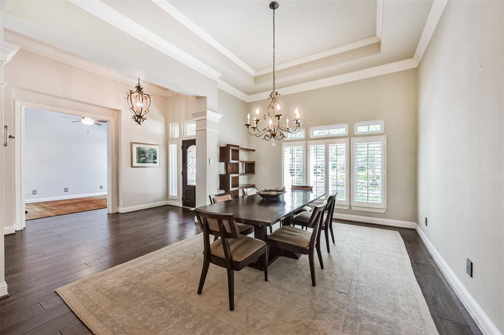 This elegant dining room features a tray ceiling, chandelier lighting, and large windows with plantation shutters. It opens into a spacious area with hardwood flooring, perfect for entertaining.