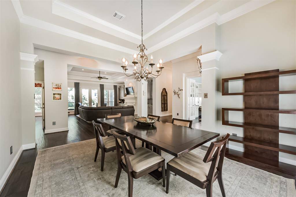 This photo showcases a spacious, open-concept dining area with elegant tray ceilings and a chandelier. It features a dark wood dining table and chairs, built-in shelving, and leads into a cozy living space with large windows providing ample natural light.