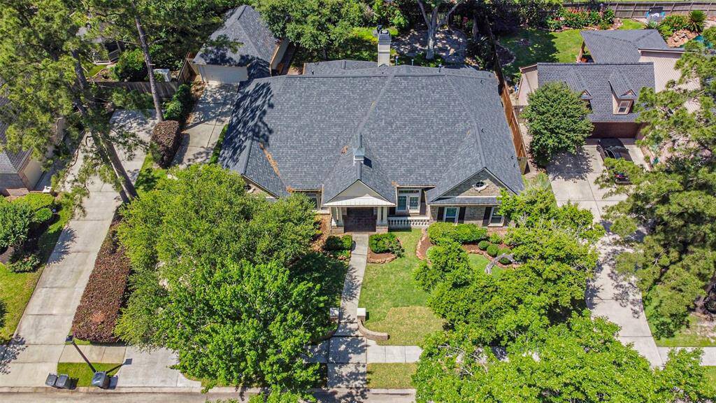 This aerial view showcases a charming single-story home with a dark roof, nestled among lush trees and well-maintained landscaping. The property features a spacious driveway and a cozy front porch, offering a welcoming curb appeal in a tranquil neighborhood setting.