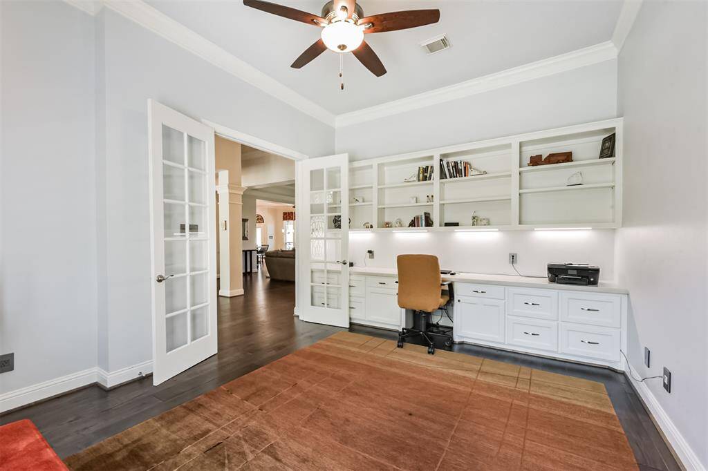 This photo showcases a bright home office with French doors and a ceiling fan. It features built-in shelves and a long desk with ample storage. The room has a neutral color palette and overlooks a spacious living area with hardwood flooring.