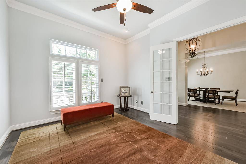 Bright room with large windows and plantation shutters, featuring a ceiling fan and wood flooring. The space opens to a dining area through French doors, creating a versatile living space.