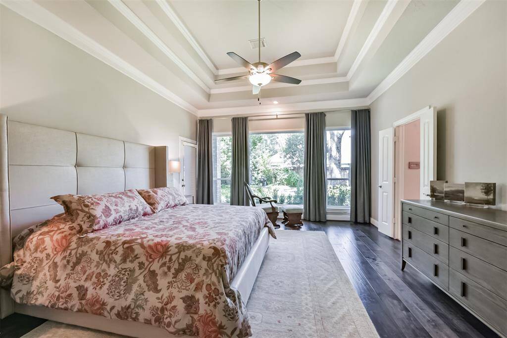 This spacious bedroom features a tray ceiling, large windows with garden views, and elegant dark wood flooring. It includes a cozy king-sized bed with floral bedding, ample natural light, and a modern ceiling fan for comfort. The room also has a sleek dresser and access to an adjoining room.
