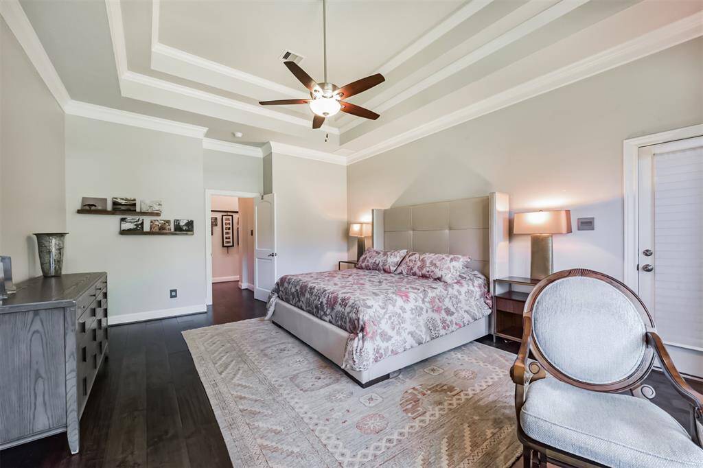 This spacious bedroom features a tray ceiling with a fan, a large bed with a cushioned headboard, and elegant decor. It includes hardwood floors, a door leading outside, and ample natural light.