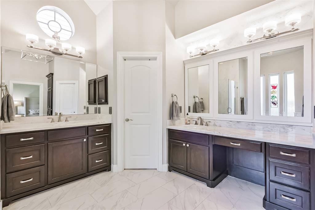 This bathroom features dual vanities with dark wood cabinets, marble countertops, and elegant lighting fixtures. The high ceiling with a unique window adds natural light, complementing the spacious feel and modern design.