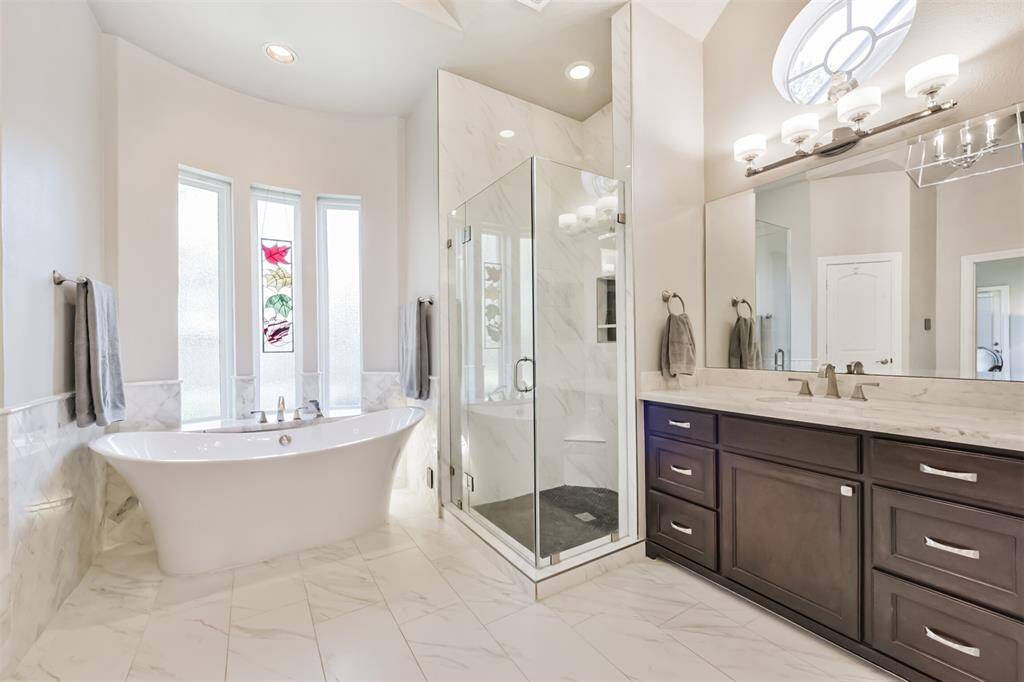 This elegant bathroom features a freestanding bathtub, a glass-enclosed shower, and a double vanity with dark wood cabinets. Natural light streams through the decorative windows, highlighting the sleek marble flooring and modern fixtures.