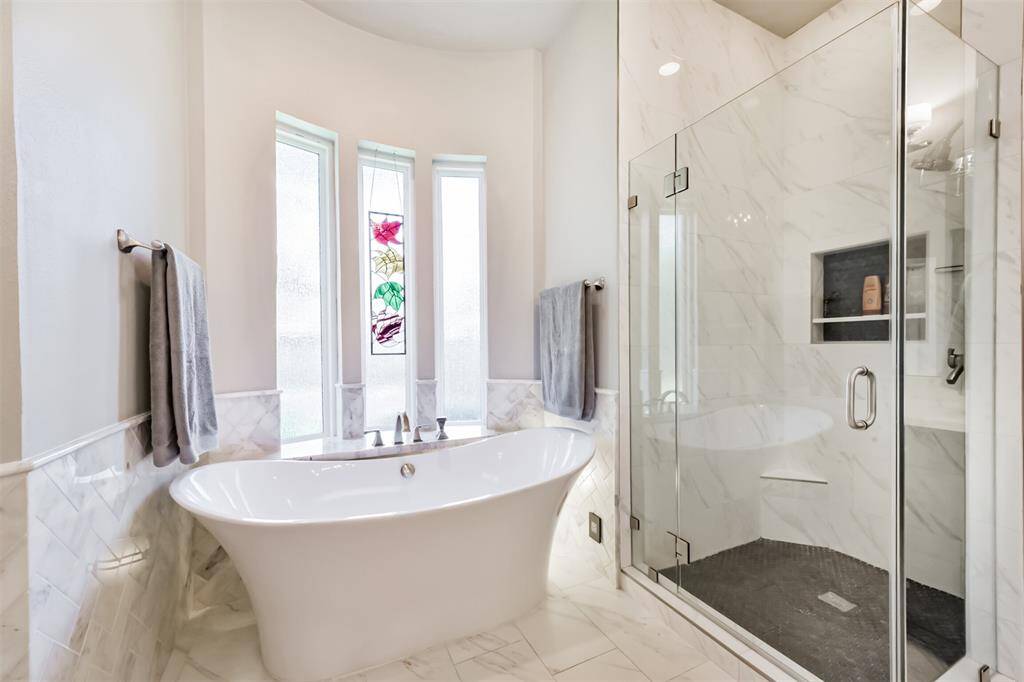 This elegant bathroom features a freestanding tub with adjacent stained glass windows, a spacious glass-enclosed shower, and sleek marble flooring and walls, offering a luxurious and serene atmosphere.