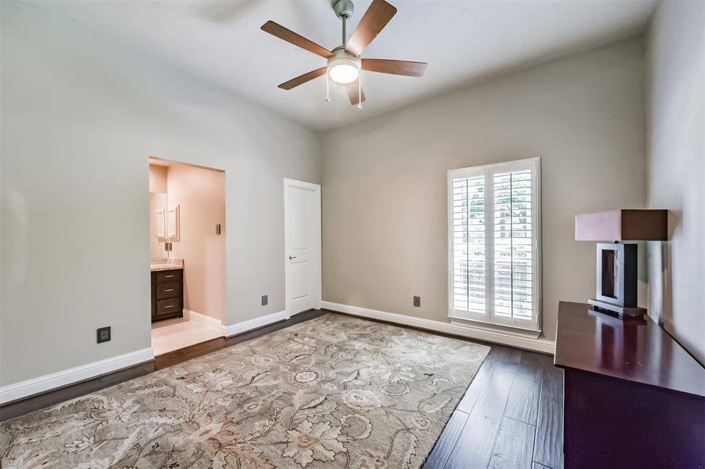 This room features hardwood flooring, a ceiling fan, and large windows with plantation shutters for ample natural light. It has neutral walls and a patterned area rug, with an adjacent bathroom visible through an open doorway.