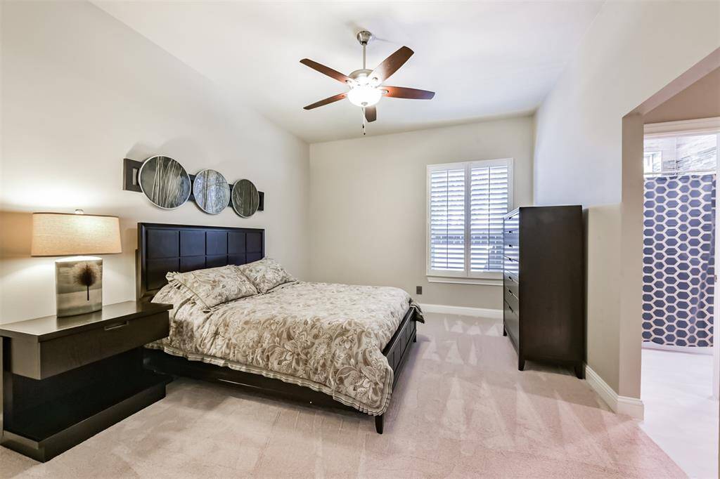 This bedroom features a cozy setup with neutral tones, a ceiling fan, and a large window with shutters. It includes a stylish bed with a patterned comforter, matching nightstand and dresser, and modern wall decor, providing a comfortable and inviting atmosphere.