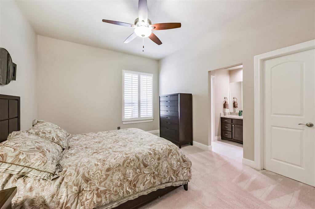 This cozy bedroom features neutral tones, a ceiling fan, and a large window with shutters for natural light. It includes direct access to an en-suite bathroom, enhancing convenience and privacy.