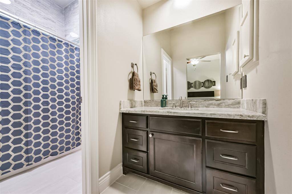This bathroom features a modern vanity with a marble countertop and dark wood cabinetry. The large mirror above enhances the space, while the shower includes a stylish hexagonal patterned curtain. Neutral walls and tile flooring complete the clean, contemporary look.