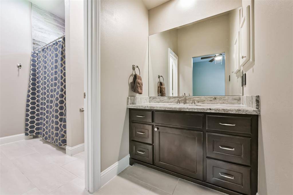 This bathroom features a modern vanity with a marble countertop, a large mirror, and ample storage. It includes a shower with a stylish geometric curtain and neutral tile flooring.