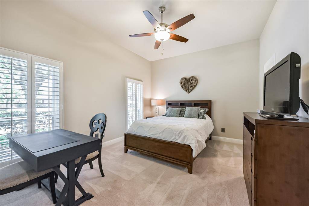 This cozy bedroom features a comfortable bed, a ceiling fan, and ample natural light from large windows with shutters. It includes a small desk and chair, and a dresser with a TV, creating a functional and inviting space.
