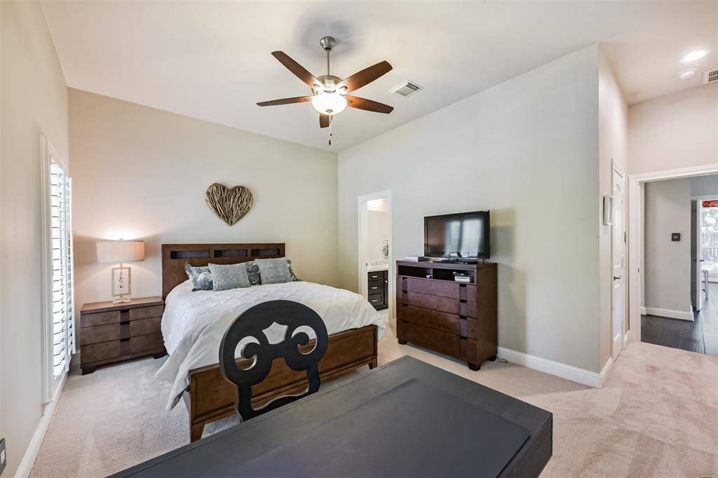This bedroom features a cozy and spacious layout with neutral walls and carpet. It includes a ceiling fan, large bed with wooden headboard, matching nightstands, and a dresser with a TV. Natural light filters through window shutters, creating a bright and inviting atmosphere.