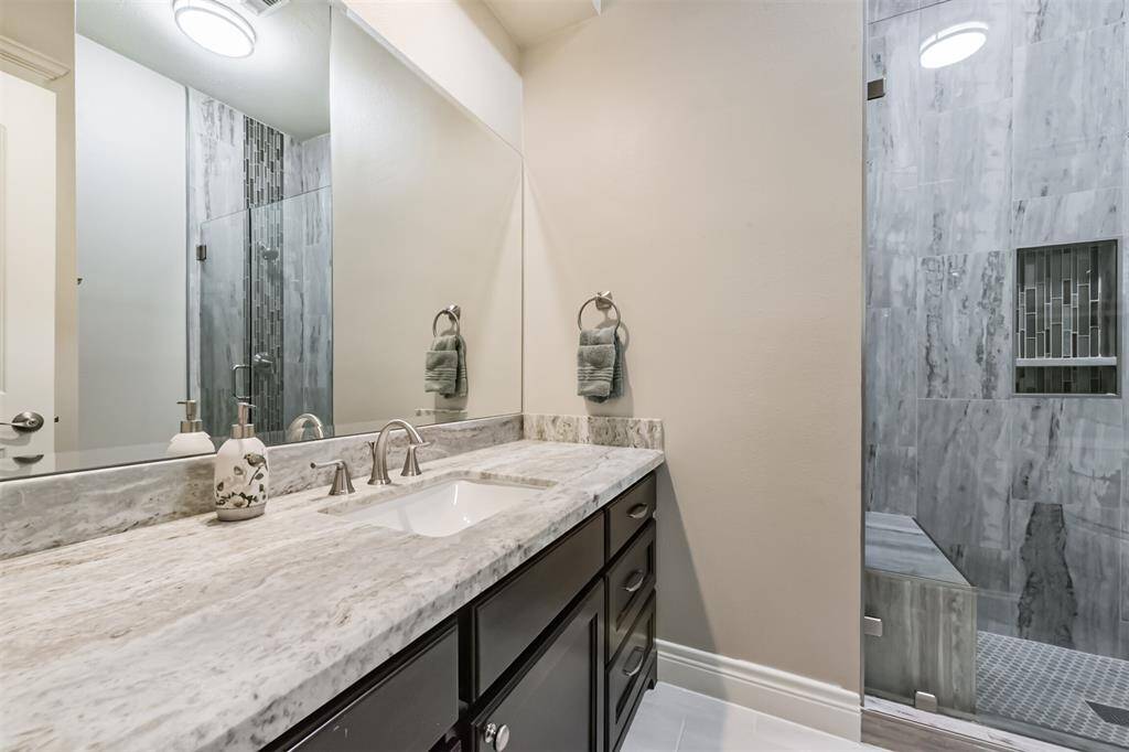 This modern bathroom features a sleek granite countertop with a single sink, elegant fixtures, and a spacious glass-enclosed shower with stylish gray tiling.