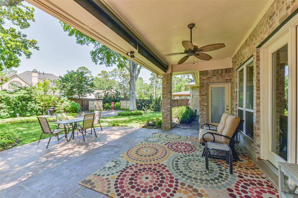 This photo features a spacious, covered patio with a ceiling fan, comfortable seating, and a colorful rug. It overlooks a well-maintained backyard with a dining set, lush greenery, and mature trees, offering a perfect space for relaxation and outdoor gatherings.