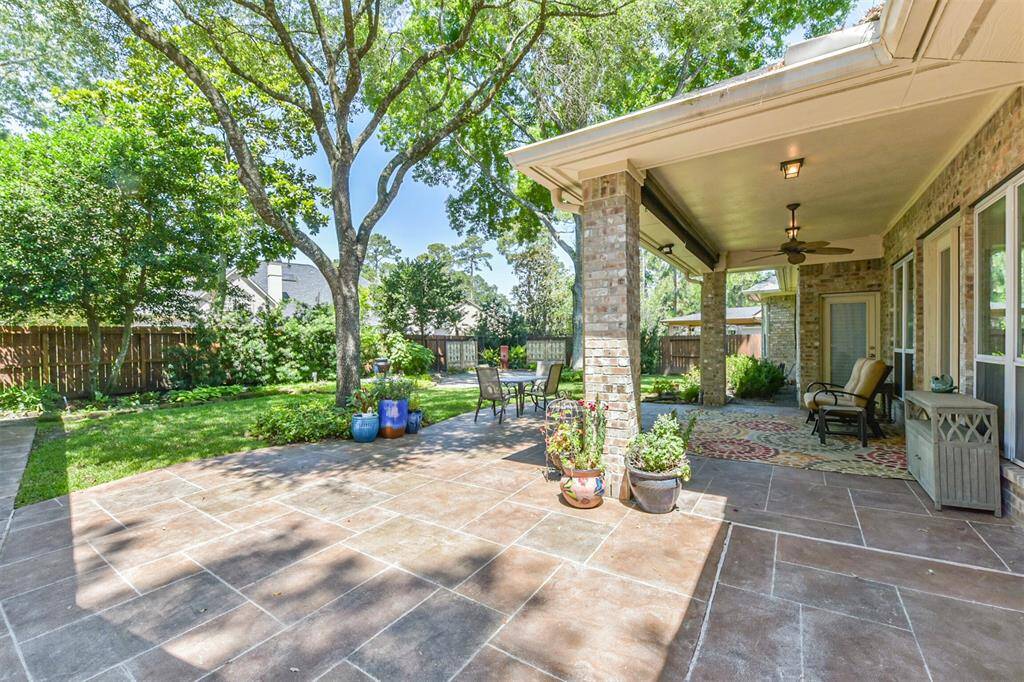 This photo showcases a spacious, shaded backyard patio with a tiled floor and brick pillars. It features a covered seating area with a ceiling fan, surrounded by lush greenery and mature trees, perfect for outdoor relaxation and entertaining.