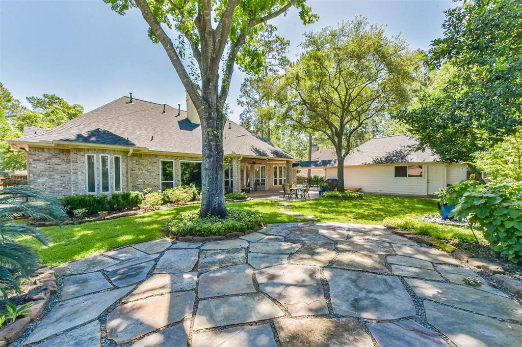 This photo showcases a charming backyard with a spacious stone patio, mature trees, and lush greenery. The brick house features large windows and a cozy outdoor seating area, perfect for relaxation and entertaining.