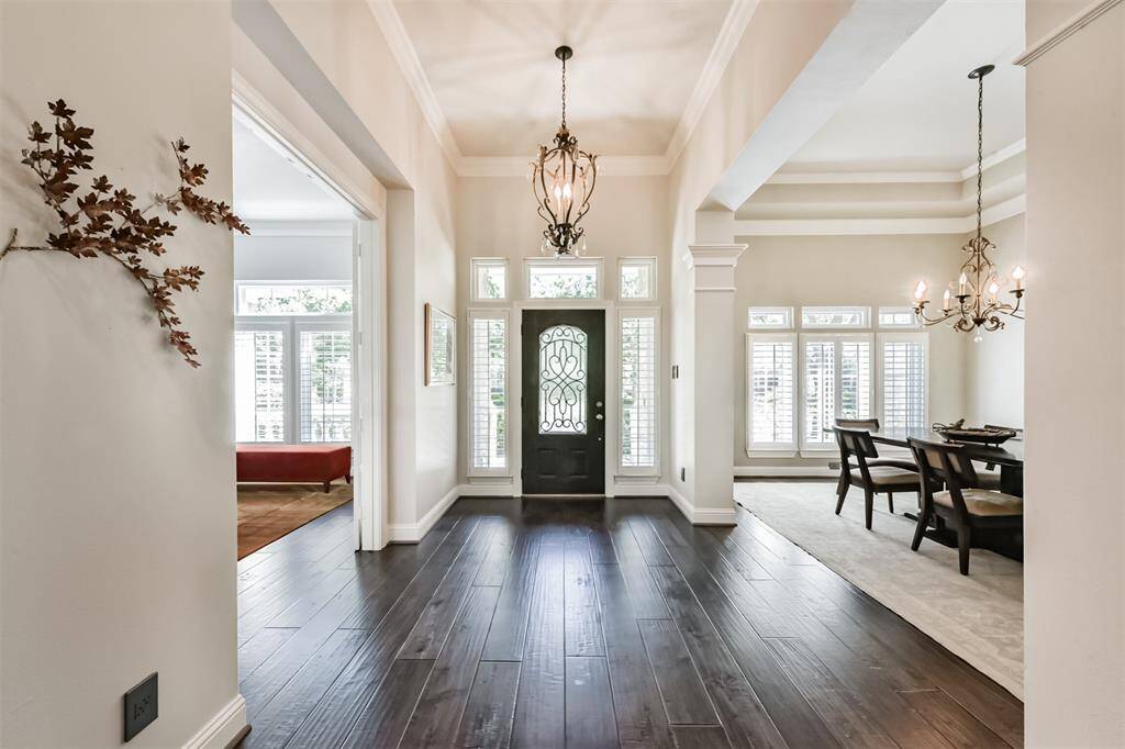 Elegant entryway with dark hardwood floors and a decorative front door flanked by tall windows. The space leads to a bright dining area with large windows and stylish chandeliers, offering a welcoming and sophisticated atmosphere.
