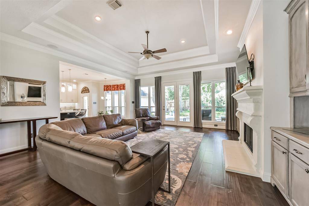 Spacious living room featuring  elegant tray ceilings, hardwood floors, and abundant natural light from large windows. It includes a cozy fireplace, comfortable leather seating, and direct access to a bright kitchen area. The room opens to a view of the outdoors, providing a seamless indoor-outdoor living experience.