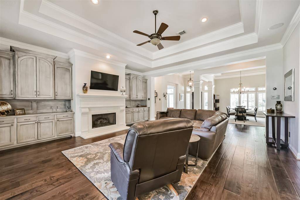 This spacious living area features elegant tray ceilings, hardwood floors, and a cozy fireplace. It has built-in cabinetry and flows seamlessly into a bright dining space with large windows. Ideal for modern living and entertaining.