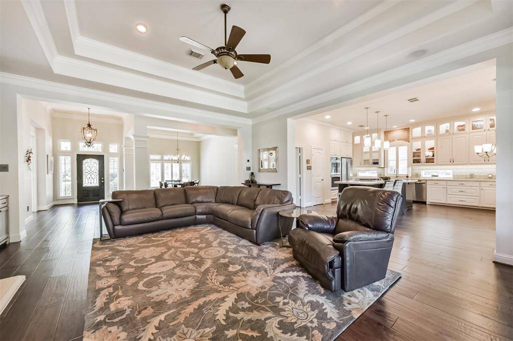 This spacious open-concept living area features elegant tray ceilings, hardwood floors, and a cozy sectional with a leather recliner. The adjoining kitchen boasts modern appliances, ample cabinetry, and a stylish island with pendant lighting. The entryway is inviting with decorative glass doors and natural light. Perfect for entertaining and family living.