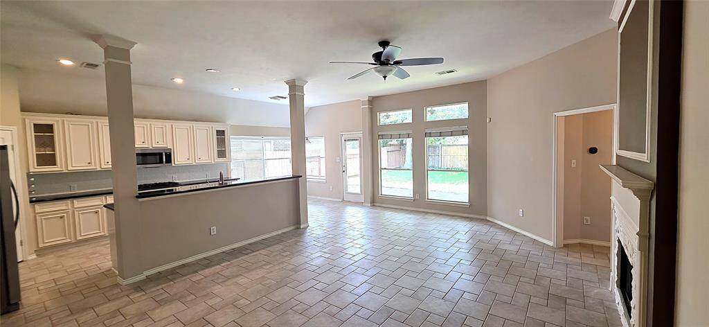 Spacious living room with large windows offering ample natural light, tiled flooring, and a stone fireplace. Neutral tones create a welcoming atmosphere. Ideal for comfortable living and entertaining.