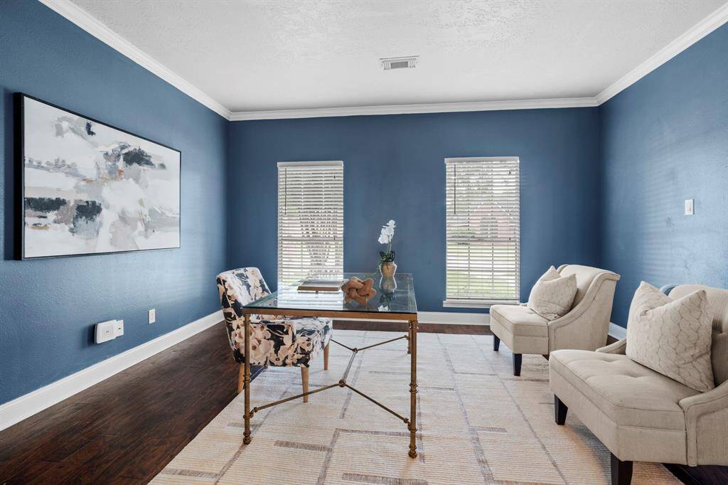 This spacious home office features rich wood-look laminate flooring, bold navy walls with crisp crown molding, and large front-facing windows that fill the room with natural light.