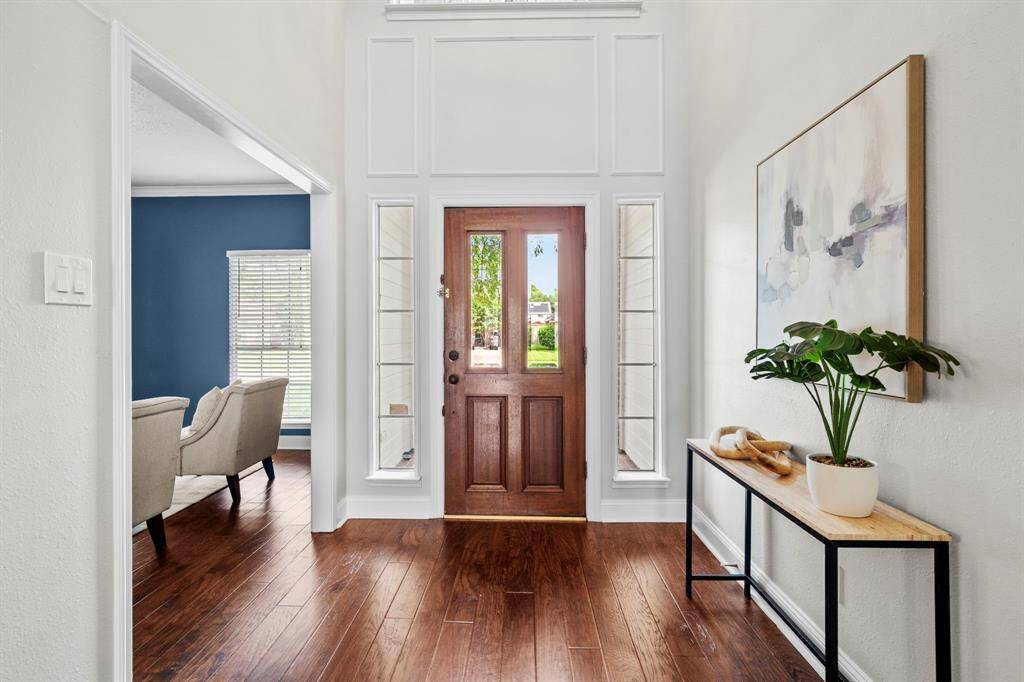 The foyer welcomes you with rich wood-look laminate flooring, tall ceilings, and classic trim details that frame the solid wood front door and side-light windows, creating a bright and inviting entrance.