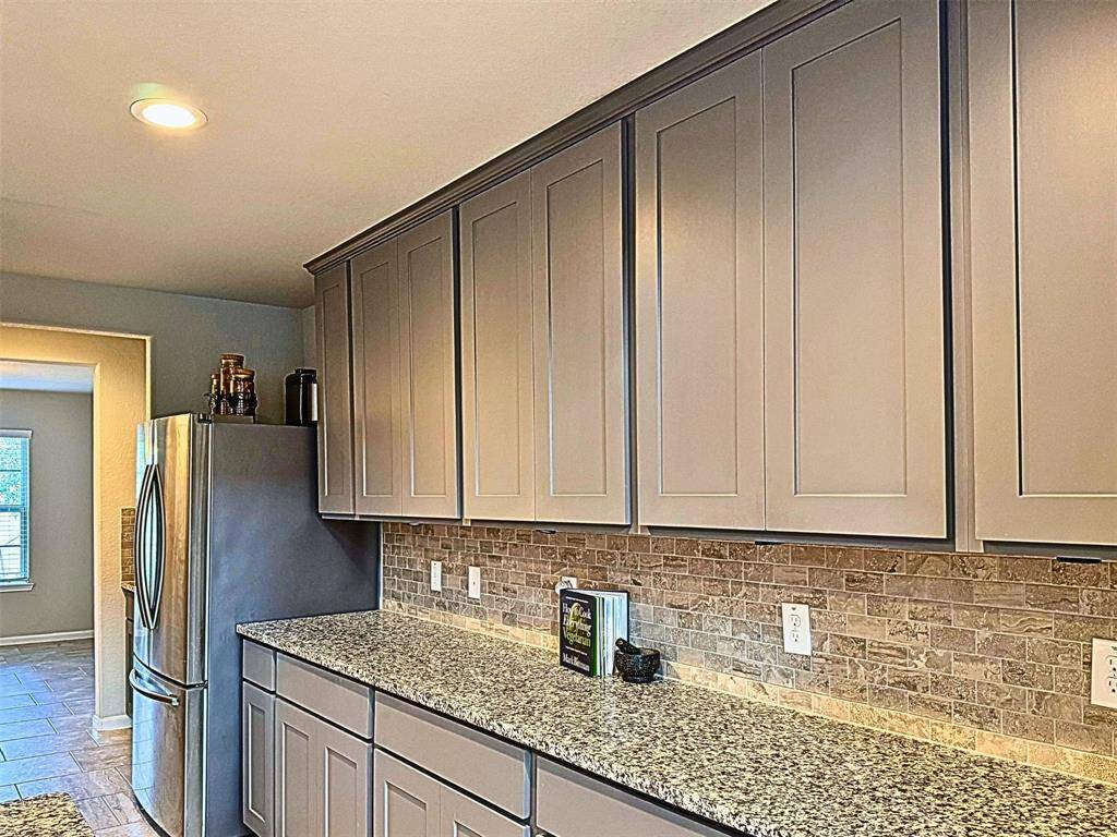 Kitchen features sleek gray cabinets, a granite countertop, and a stylish stone backsplash. It includes a stainless steel refrigerator and flows into a bright dining area with a large window, providing a modern and spacious feel.